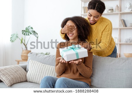 Happy celebration and congratulations from friend, idea for present and birthday. Smiling cute millennial mixed race lady closes eyes and gives gift to african american woman in living room interior