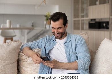 Happy Cellphone User Getting Good News, Reading Text Message On Smartphone, Smiling At Screen. Millennial Man Using Mobile Phone On Couch At Home, Shopping Online, Chatting On Social Media