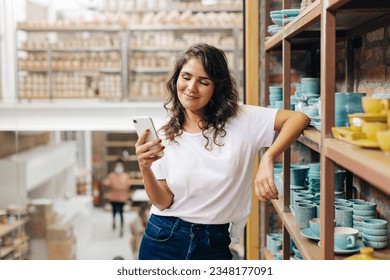 Happy ceramic store owner reading a text message on her smartphone. Businesswoman managing a store with a collection of earthenware products. Cheerful young woman running a successful small business. - Powered by Shutterstock