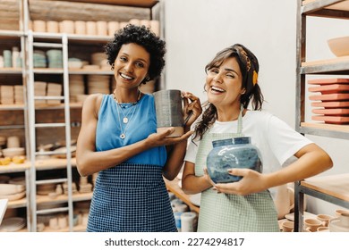 Happy ceramists smiling at the camera while holding their handmade ceramic products. Two craftswomen working together in their pottery studio. Creative young women running a successful small business.