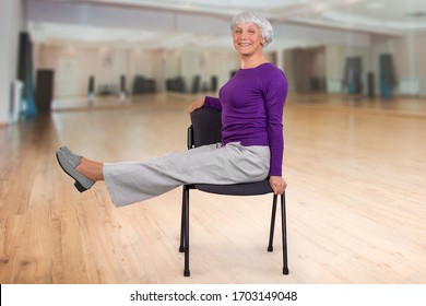 Happy charming beautiful elderly woman is doing exercise on a chair. Exercising gymnastics for health in the fitness room. Sports training.