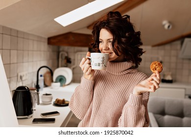 Happy charming girl with curly hair with brown hair, drinking latte and eating cookies at kitchen . High quality photo