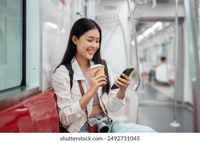 A happy, charming young Asian female tourist with a backpack is using her phone, reading text messages while taking a sky train or metro train, commuting in a city. people and transportation concepts - Powered by Shutterstock