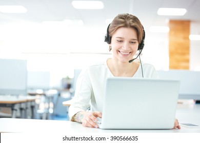 Happy Charming Young Woman Sitting And Working With Laptop Using Headset In Office