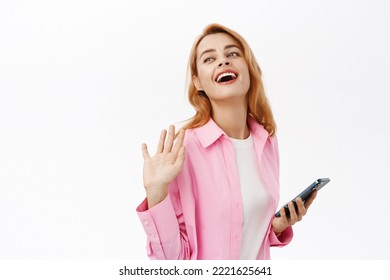 Happy chatting girl holding smartphone and saying hello to someone, waving hand, standing over white background