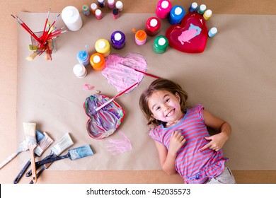 Happy Cheeky Little Girl Artist With Her Art Supplies Lying Surrounded By Colorful Jars Of Paint, Brushes And Pretty Heart Designs On A Sheet Of Brown Paper Grinning At The Camera, Overhead View