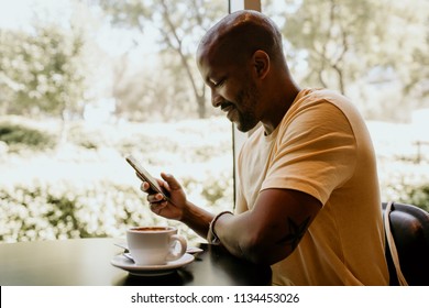 Happy cheerful African hipster holding mug, drinking fresh cappuccino, browsing internet and checking newsfeed on social media.Man using cell phone during coffee break at modern cafe
