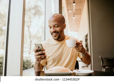 Happy cheerful African hipster holding mug, drinking fresh cappuccino, browsing internet and checking newsfeed on social media.Man using cell phone during coffee break at modern cafe - Powered by Shutterstock