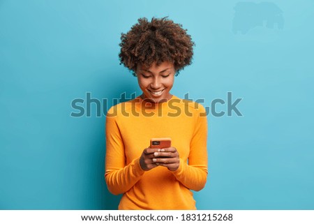 Happy cheerful Afro American woman looks at screen of smart phone enjoys online chatting types text message surfs social networks dressed casually poses against blue background. Technology concept