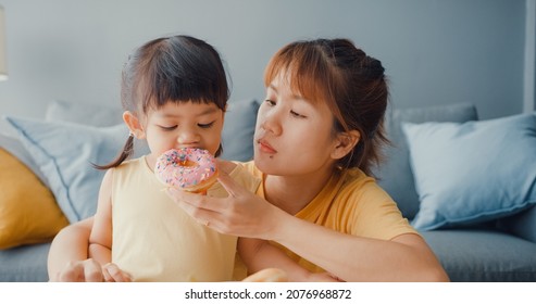 Happy Cheerful Asia Family Mom And Toddler Girl Eating Donuts And Having Fun Relax Enjoy On Couch In Living Room At House. Spending Time Together, Social Distance, Quarantine For Coronavirus.