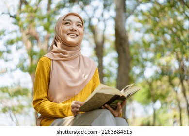 Happy cheerful Asian beautiful Muslim woman reading a book - Koran under the tree. Female muslim student studying an Islam Koran. - Powered by Shutterstock