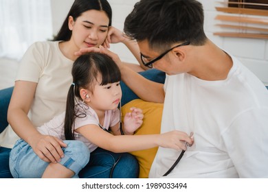 Happy Cheerful Asian Family Dad, Mom And Daughter Playing Funny Game As Doctor Having Fun On Sofa At Home. Self-isolation, Stay At Home, Social Distancing, Quarantine For Coronavirus Prevention.