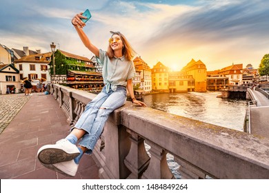 Happy and cheerful Asian girl traveler takes a selfie on the Saint Martin bridge in the Petit France area in Strasbourg. Colorful and dramatic sunset at the background - Powered by Shutterstock