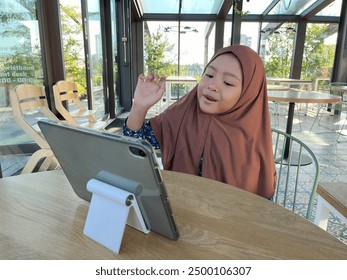 Happy cheerful Asian little Muslim girl siting and learning online with mobile tablet in cafe or restaurant. - Powered by Shutterstock