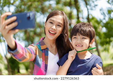 Happy cheerful Asian lovely little boy   mother sitting under the tree at park in the morning   drawing picture  Lovely mother   son taking selfie photography together 