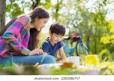 Happy cheerful Asian lovely little boy   mother sitting under the tree at park in the morning   drawing picture  Asian woman spending time in weekend and her lovely little son 