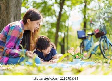 Happy cheerful Asian lovely little boy   mother sitting under the tree at park in the morning   drawing picture  Asian woman spending time in weekend and her lovely little son 