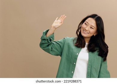 Happy cheerful Asian student young woman in khaki green shirt greet someone waving hand posing isolated on over beige pastel studio background. Cool fashion offer. Lifestyle and Emotions concept