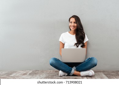 Fröhliche asiatische Frau, die am Laptop arbeitet, während sie auf dem Boden sitzt, mit Beinen einzeln auf grauem Hintergrund gekreuzt