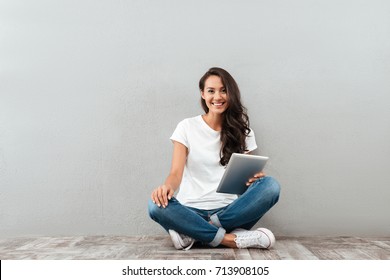 Fröhliche asiatische Frau, die am Tablet-Computer arbeitet, während sie einzeln auf grauem Hintergrund sitzt