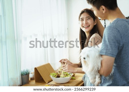 Happy cheerful Asian young couple preparing a vegetable salad in kitchen together in weekend, man holding lovely cute Maltese dog to see his girlfriend making a salad in kitchen.