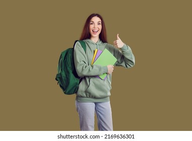 Happy cheerful beautiful college or university student girl in green hoodie with backpack standing isolated on solid greenish brown color background, holding her books, showing thumbs up and smiling