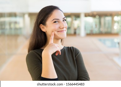 Happy Cheerful Beautiful Woman Touching Face With Index Finger. Young Woman In Casual Posing Indoors With Glass Wall Interior In Background. Skin Care Or Female Portrait Concept