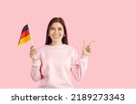 Happy cheerful beautiful young German woman in casual sweatshirt standing isolated on pastel pink background, holding flag of Germany, pointing her index finger at copy space on the right, and