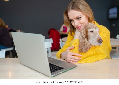 Happy cheerful blonde woamn hugs her funny cocker spaniel dog at the table with laptop and posing for camera.