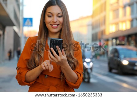 Happy cheerful casual woman typing with finger on smart phone on city street in spring at sunset