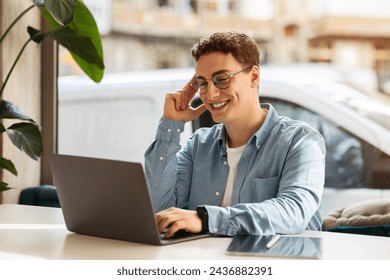 A happy cheerful caucasian young man with curly hair and glasses smiling at his laptop in a bright room with a plant and a white van visible through the window. Work, study remotely, freelance - Powered by Shutterstock