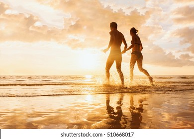 Happy cheerful couple having fun running to the sea together and doing splashes of water on a tropical beach at sunset - concept about romantic vacation, honeymoon
