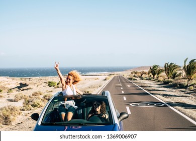 Happy Cheerful Couple Of Young Caucasian Crazy Woman With Curly Hair Enjoy The Travel And Vacation Staying Outside The Roof Of A Convertible Car With Long Road And Ocean In Background - Happiness Joy