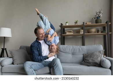 Happy cheerful dad holding excited funny preschool son kid in arms upside down, sitting on couch, laughing, having fun, playing active games with child, enjoying family leisure time, fatherhood