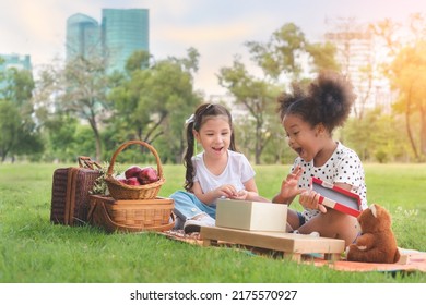 Happy Cheerful Ethnic Girls Open Surprise Puzzle Gift Box Together Between Picnic At Outdoors Park , Relationship Little Kids, Diverse Ethnic Concept, White And Black Together Concept,