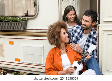 Happy cheerful family of tree parents and kid daughter singing playing ukulele guitar while traveling by trailer van motor home. Adventures ahead, away from everything - Powered by Shutterstock