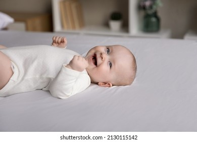 Happy Cheerful Few Month Baby Resting On White Linen, Lying Back On Pale Sheet, Looking Away, Smiling, Laughing, Feeling Joy. Infant Boy Or Girl In White Bodysuit Relaxing On Soft Mattress. Childbirth