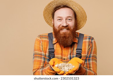 Happy cheerful fun young bearded man wear straw hat overalls work in garden hold in palms pumpkin seeds look camera isolated on plain pastel light beige color background studio. Plant caring concept - Powered by Shutterstock