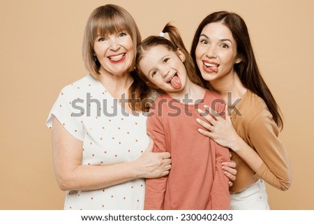 Happy cheerful funny women wear casual clothes with child kid girl 6-7 years old. Granny mother daughter show tongue kidding look camera isolated on plain beige background. Family parent day concept