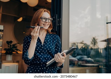 Happy cheerful ginger business woman freelancer with laptop holds mobile phone and talks on speakerphone with friend or client, uses virtual assistant or records audio message on smartphone outdoors - Powered by Shutterstock