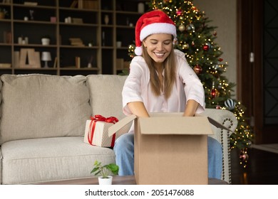 Happy Cheerful Girl In Christmas Santa Hat Receiving Delivery Box With Gifts, Producing Wrap From Package, Unpacking Parcel On Couch At Home Xmas Tree Lights. Shipping, Courier Service On Holidays