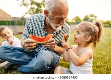 Happy cheerful granddad is sitting in backyard and eating watermelon slices with grandchildren while enjoying summertime. - Powered by Shutterstock