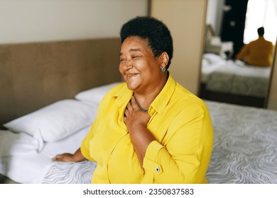 Happy cheerful grateful african american grandmother sitting on bed in yellow shirt keeping hand on chest smiling, being thankful for best life, thinking about her past, funny unforgettable moments - Powered by Shutterstock