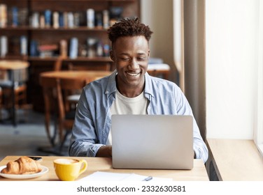 Happy cheerful handsome young black guy in casual outfit working online on laptop computer at cozy city cafe, entrepreneur responding emails while have breakfast at restaurant, copy space - Powered by Shutterstock