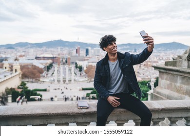 Happy Cheerful Latino Male Traveller Dressed In Stylish Outfit Resting During Spanish Vacations To Barcelona City Clicking Selfie Pictures On Montjuic Area, Millennial Man Photographing Himself