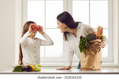 Happy, Cheerful Little Daughter Makes A Crazy, Funny Face Covering Her Eyes With Two Red Tomatoes. Family Having Fun While Unpacking Natural Vegetarian Foods From Paper Grocery Bags In The Kitchen