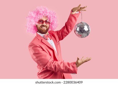 Happy cheerful man with ginger beard, wearing pink outfit, bow tie, funny curly clown wig and cool disco glasses standing on pink studio background, smiling and holding mirror ball. Fun party concept