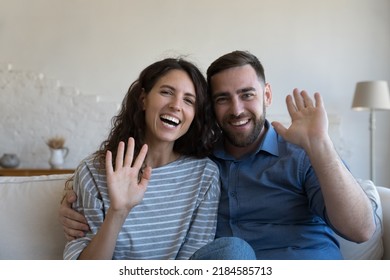 Happy Cheerful Married Couple Video Call Shot Portrait. Dating Man And Woman In Love Sitting On Home Couch, Hugging, Waving Hand Hello Camera With Toothy Smiles, Saying Hi, Talking, Laughing