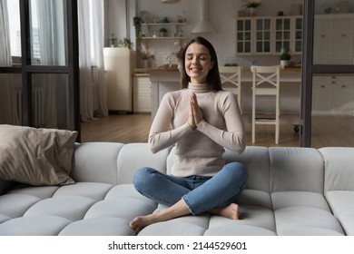 Happy cheerful meditating girl practicing on couch at home, sitting in sukhasana, keeping Namaste hands, closed eye, smiling, doing yoga. Mindfulness, stress relief concept