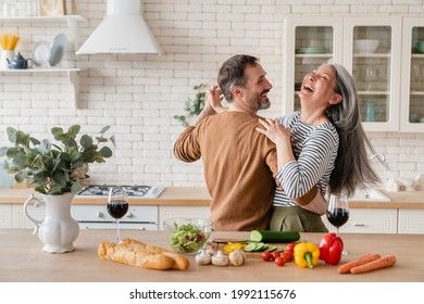 Happy cheerful middle-aged mature couple family parents dancing together in the kitchen, preparing cooking food meal for romantic dinner, spending time together. Active seniors